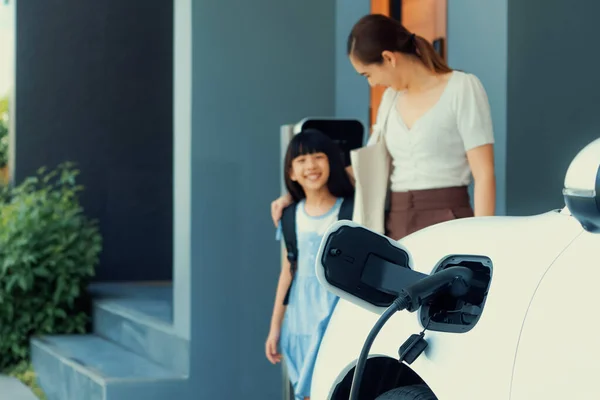 Ev充電器に接続された家庭用充電ステーションで充電する電気自動車に焦点を当て 進歩的な母親と娘がエネルギーの持続可能性の概念として歩く背景をぼかしています — ストック写真