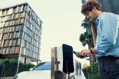 Takım elbiseli ilerici iş adamı şarj kablosu ve yenilenebilir enerji enerjili bir elektrikli araçla şarj istasyonunda dururken telefonundan EV araba aküsünün durumuna bak..