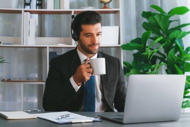 Çağrı merkezi operatörünün müdürü bilgisayarla uğraşırken masasında kahvesiyle oturuyor. Kulaklık takan ve siyah takım giyen bir ofis çalışanı müşteri desteği ya da tele pazarlama üzerinde çalışıyor. coşkulu