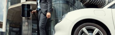 Below view of progressive businessman with electric car recharging at public charging station at modern city residential building. Eco friendly rechargeable EV car powered by alternative clean energy.