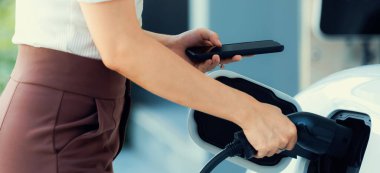 İlerici kadın elektrikli arabasının fişini şarj istasyonuna takıyor. İlerici bir yaşam tarzında elektrikli araç kullanımı kavramı temiz çevreye katkıda bulunur.