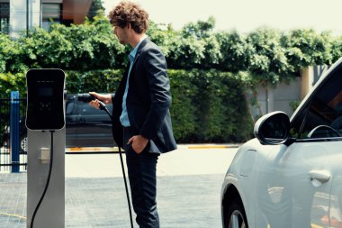 Modern şehir yerleşim bölgesindeki kamu şarj istasyonunda elektrikli araba şarj eden yenilikçi bir işadamı. Çevre dostu şarj edilebilir araba alternatif temiz enerjiyle çalışıyor..