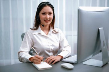 Kulaklık ve mikrofonlu Asyalı çağrı merkezi çalışma alanı üzerinde çalışıyor ve kameraya bakıyor. Kadın operatör istisnai müşteri hizmetleri sunar. Yardımcı çağrı merkezi ajan portresi. Coşkulu.