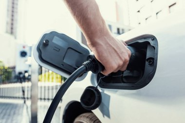 Closeup progressive man holding EV charger plug from public charging station for electric vehicle with background of residential building as concept eco-friendly sustainability energy car concept.
