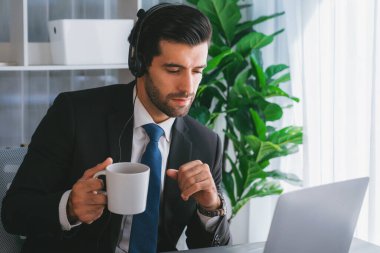 Çağrı merkezi operatörünün müdürü bilgisayarla uğraşırken masasında kahvesiyle oturuyor. Kulaklık takan ve siyah takım giyen bir ofis çalışanı müşteri desteği ya da tele pazarlama üzerinde çalışıyor. coşkulu