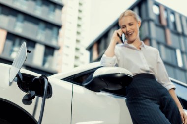 Focus charging-electric car with EV charger at charging station with blur businesswoman talking on phone with residential building in background as progressive lifestyle concept.