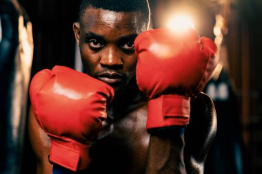 Boksör üstsüz, Afro-Amerikan siyah boksör savunma pozisyonu almış kırmızı eldiven giymiş, arka planda boks ekipmanları ve kum torbalarıyla spor salonunda dövüşmeye ve yumruk atmaya hazır. Impetus