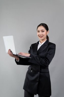 Kendine güvenen genç iş kadını izole edilmiş bir zeminde duruyor ve resmi siyah takım elbiseyle dizüstü bilgisayarda çalışıyor. Ofisteki kadın ya da yönetici, zeki ve profesyonel görünümlü. Coşkulu.