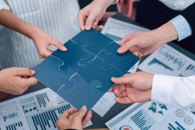 Closeup top view business team of office worker putting jigsaw puzzle together over table filled with financial report paper in workplace with manager to promote harmony concept in meeting room.