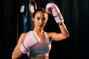 Asya 'lı kadın Muay Thai boksörü kamera önünde boksör yumruğu. Arka planda boks ekipmanıyla spor salonunda poz vermeye hazır. Kararlı gözler ve meydan okumaya hazır olun. Impetus