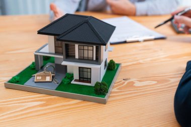Ev modeli ahşap toplantı masasında, emlakçı ve müşterinin bulanık geçmişinde ev kredisi veya kira kontratının şartlarını tartışırken görüntülenir. Varlık