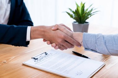 İki iş adamı, şirket ofisinde yasal belge ve kalemle imzalanan sözleşmenin ardından iş ortaklığı ve işbirliğini simgeleyen tokalaşıyor. Varlık