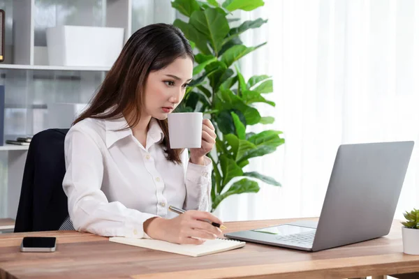ラップトップを使用してコーヒーを飲みながら作業する現代のオフィスデスクで若いアジアの熱狂的なビジネス女性 勤勉で魅力的なオフィスの女性は彼女のオフィスワークスペースでコンピュータノートブックに取り組んで — ストック写真