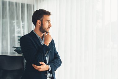 Siyah takım elbiseli yakışıklı iş adamı, modern ofis portresine güvenerek duruyor. Düşünceli bir ifadeyle iş hakkında derin düşüncelere dalmış bir sonraki hamlesini stratejik olarak düşünüyor. Varlık