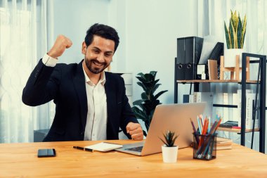 Siyah takım elbise giymiş heyecanlı ve mutlu bir iş adamı başarılı bir iş tanıtımı, enerji ve neşe kazandıktan sonra ofis masasında kutlama jesti olarak elini kaldırır. Coşkulu