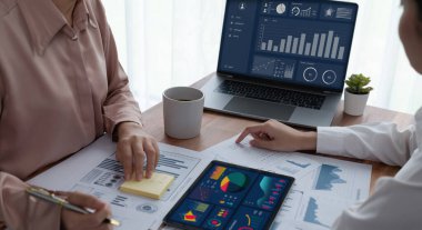 Analist ekip arkadaşı dijital gösterge panelinde finansal verileri tartışır, grafikleri dizüstü bilgisayar ve tablet ekranda analiz eder. Modern ofis pazarlama planlamak için iş zekasını kullanır. Coşkulu.