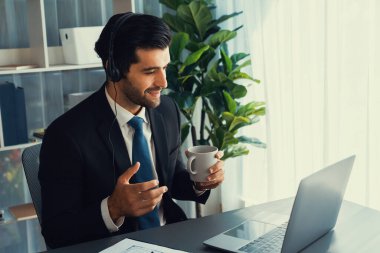 Çağrı merkezi operatörünün müdürü bilgisayarla uğraşırken masasında kahvesiyle oturuyor. Kulaklık takan ve siyah takım giyen bir ofis çalışanı müşteri desteği ya da tele pazarlama üzerinde çalışıyor. coşkulu
