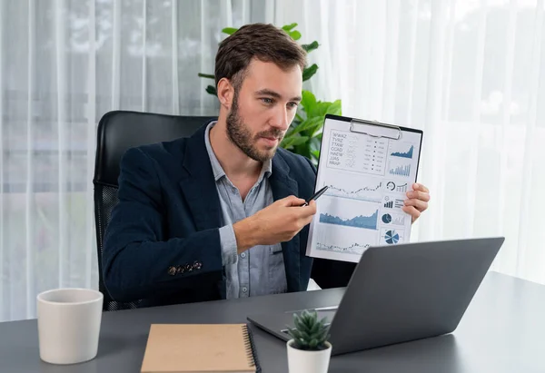 Professional businessman in black suit present financial data or BI paper clipboard to laptop camera during online presentation meeting. Remote work with virtual meeting presentation in office. Entity