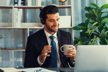 Çağrı merkezi operatörünün müdürü bilgisayarla uğraşırken masasında kahvesiyle oturuyor. Kulaklık takan ve siyah takım giyen bir ofis çalışanı müşteri desteği ya da tele pazarlama üzerinde çalışıyor. coşkulu