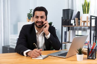 Çalışkan işadamı, ofis masasında dizüstü bilgisayarla çalışırken müşterileriyle telefonda konuşmakla meşgul. Cep telefonlu, modern, çalışkan ofis çalışanı yaşam tarzı kavramı olarak. Coşkulu