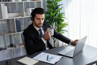 Çağrı merkezi operatörünün müdürü bilgisayarla uğraşırken masasında kahvesiyle oturuyor. Kulaklık takan ve siyah takım giyen bir ofis çalışanı müşteri desteği ya da tele pazarlama üzerinde çalışıyor. coşkulu