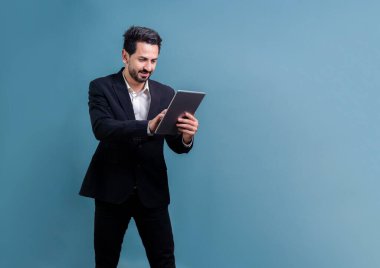 Resmi takım elbiseli kendine güvenen işadamı elinde tabletle sürpriz bir şekilde terfi ya da reklam arıyor. Yüz ifadeleri ve jestler, izole bir arkaplanda heyecan ve şaşkınlığa işaret ediyor. Coşkulu