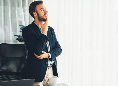 Siyah takım elbiseli yakışıklı iş adamı, modern ofis portresine güvenerek duruyor. Düşünceli bir ifadeyle iş hakkında derin düşüncelere dalmış bir sonraki hamlesini stratejik olarak düşünüyor. Varlık