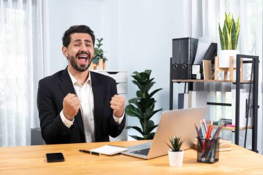 Siyah takım elbise giymiş heyecanlı ve mutlu bir iş adamı başarılı bir iş tanıtımı, enerji ve neşe kazandıktan sonra ofis masasında kutlama jesti olarak elini kaldırır. Coşkulu