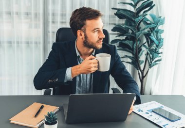 İşadamı laptopuyla çalışıyor ve çalışma masasında kahve içiyor. Akıllı yönetici şirket bilgisayarındaki finansal verileri ve pazarlama planını inceledikten sonra kahve molası veriyor. Varlık