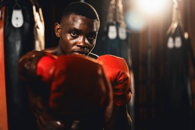 Üstsüz boksör pozu veren Afro-Amerikan Siyah Boksör yumruğunu agresif bir şekilde yumrukladı ve arka planda kum torbası ve boks ekipmanıyla spor salonunda dövüşmeye hazırdı. Impetus