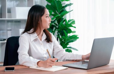 Modern ofis masasındaki Asyalı hevesli genç iş kadını çalışmak ve notlar yazmak için dizüstü bilgisayar kullanıyor. Çalışkan ve çekici ofis hanımı ofisinde bilgisayar defteri üzerinde çalışıyor..