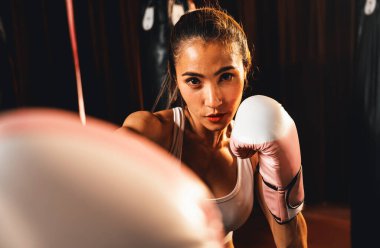Asya 'lı kadın Muay Thai boksörü kamera önünde boksör yumruğu. Arka planda boks ekipmanıyla spor salonunda poz vermeye hazır. Kararlı gözler ve meydan okumaya hazır olun. Impetus