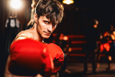 Üstsüz boksör pozu veren beyaz boksör, kameranın önünde agresif bir duruşta yumruğunu yumrukluyor ve arka planda kum torbası ve boks ekipmanıyla spor salonunda dövüşmeye hazır. Impetus