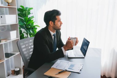 Modern ofis masasındaki modern profesyonel iş adamı elinde kahveyle çalışmak için dizüstü bilgisayar kullanıyor. Ofisinde bilgisayar defteri üzerinde çalışan çalışkan bir ofis çalışanı. coşkulu