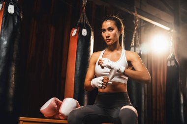 Kararlı Asyalı kadın Muay Thai boksörü kaslı, fiziksel olarak hazır ya da boks eldiveni giymiş, ya da boks eldiveni giymiş, spor salonundaki ringde yoğun boks antrenmanına hazırlanıyor. Impetus
