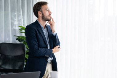 Siyah takım elbiseli yakışıklı iş adamı, modern ofis portresine güvenerek duruyor. Düşünceli bir ifadeyle iş hakkında derin düşüncelere dalmış bir sonraki hamlesini stratejik olarak düşünüyor. Varlık