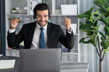 Siyah takım elbiseli heyecanlı ve mutlu iş adamı, ofis masasında kutlama jesti olarak kolunu kaldırdı. İş yerinde başarılı bir çağrı merkezi ya da telesales operatörü kutlama yapıyor. Coşkulu