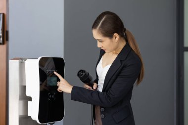 Asian woman holding and pointing an EV plug, a home charging station providing a sustainable power source for electric vehicles. Alternative energy for progressive lifestyle.
