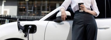 Businesswoman drinking coffee, leaning on electric vehicle recharging at public charging station with residential apartment condos building in background as progressive lifestyle by eco-friendly car.