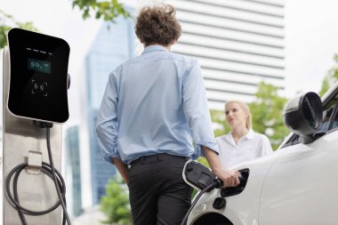 Progressive businessman and businesswoman with electric car parking and connected to public charging station before driving around city center. Eco friendly rechargeable car powered by clean energy.