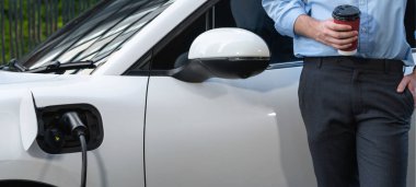 Progressive eco-friendly concept of parking EV car at public electric-powered charging station in city with blur background of businessman leaning on recharging-electric vehicle with coffee.