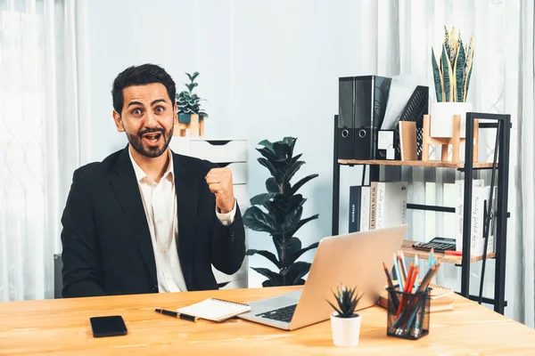 stock image Excited and happy businessman dressed in black formal suit, raise his arm in celebratory gesture at his office desk, after successful job promotion, energy and joy as business winner. Fervent