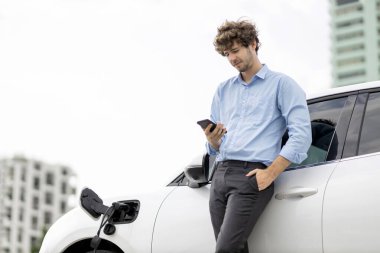 Progressive businessman talking on the phone, leaning on electric car recharging with public EV charging station, apartment condo residential building on the background as green city lifestyle.