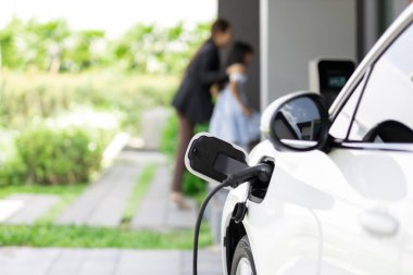 Focus EV car recharging at home charging station with blurred progressive woman and young girl in background for alternative clean energy technology concept for renewable electric vehicle.