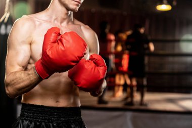 Boksör gömleksiz, beyaz erkek boksör savunma pozisyonu almış kırmızı eldiven giymiş arka planda ring ve boks ekipmanıyla dövüşmeye ve yumruk atmaya hazır. Impetus
