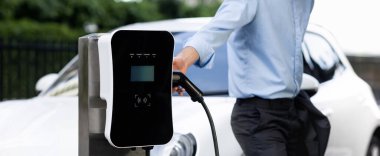 Closeup progressive suit-clad businessman with his electric vehicle recharge his car on public charging station in modern city with power cable plug and renewable energy-powered electric vehicle.