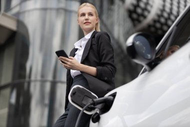 Businesswoman wearing black suit using smartphone, leaning on electric car recharge battery at charging station in city residential building with condos and apartment. Progressive lifestyle concept.