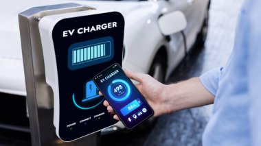 Akıllı telefon ekran bataryasını elinde tutan iş adamı akıllı EV mobil uygulama ile otoparktaki şarj istasyonundan elektrik şarj eden EV araba. İnceleyin