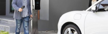 Closeup progressive asian man install cable plug to his electric car with home charging station. Concept of the use of electric vehicles in a progressive lifestyle contributes to clean environment.