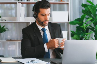 Çağrı merkezi operatörünün müdürü bilgisayarla uğraşırken masasında kahvesiyle oturuyor. Kulaklık takan ve siyah takım giyen bir ofis çalışanı müşteri desteği ya da tele pazarlama üzerinde çalışıyor. coşkulu